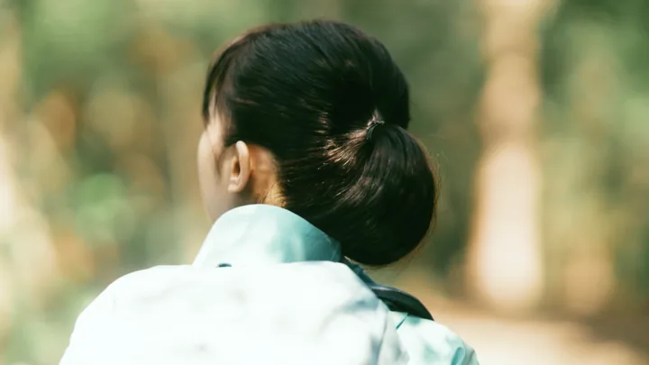 A young woman in a teal jacket seen from behind in a forest