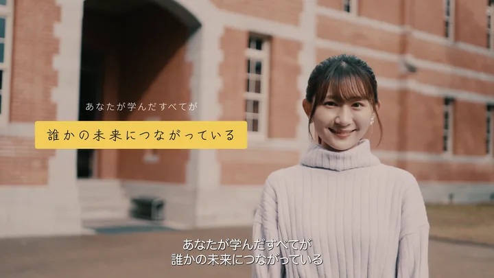 A student smiling in front of the red-brick building — 「あなたが学んだすべてが誰かの未来につながっている」