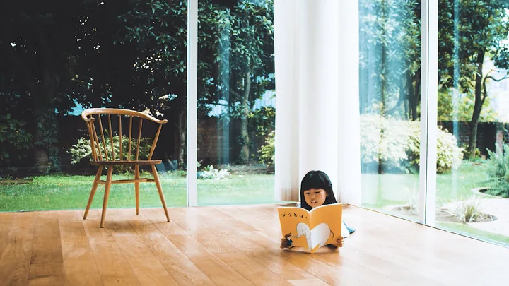 A girl reading a picture book on a wooden floor by a garden window