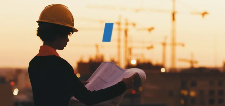 An engineer in a hard hat reading plans at a construction site at sunset