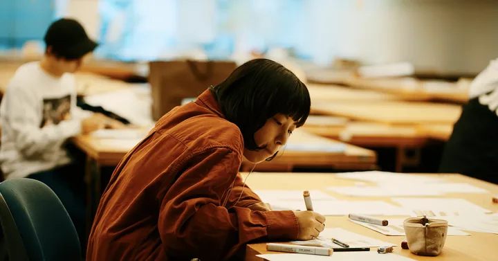 A student in an orange jacket drawing at a desk